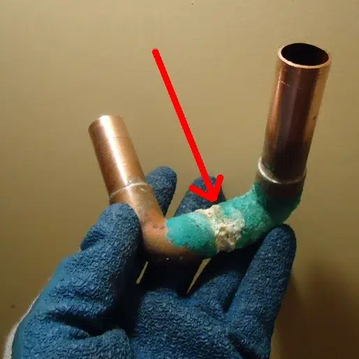 Section of copper pipe with a pinhole leak, green corrosion and a white crust, held in a gloved hand